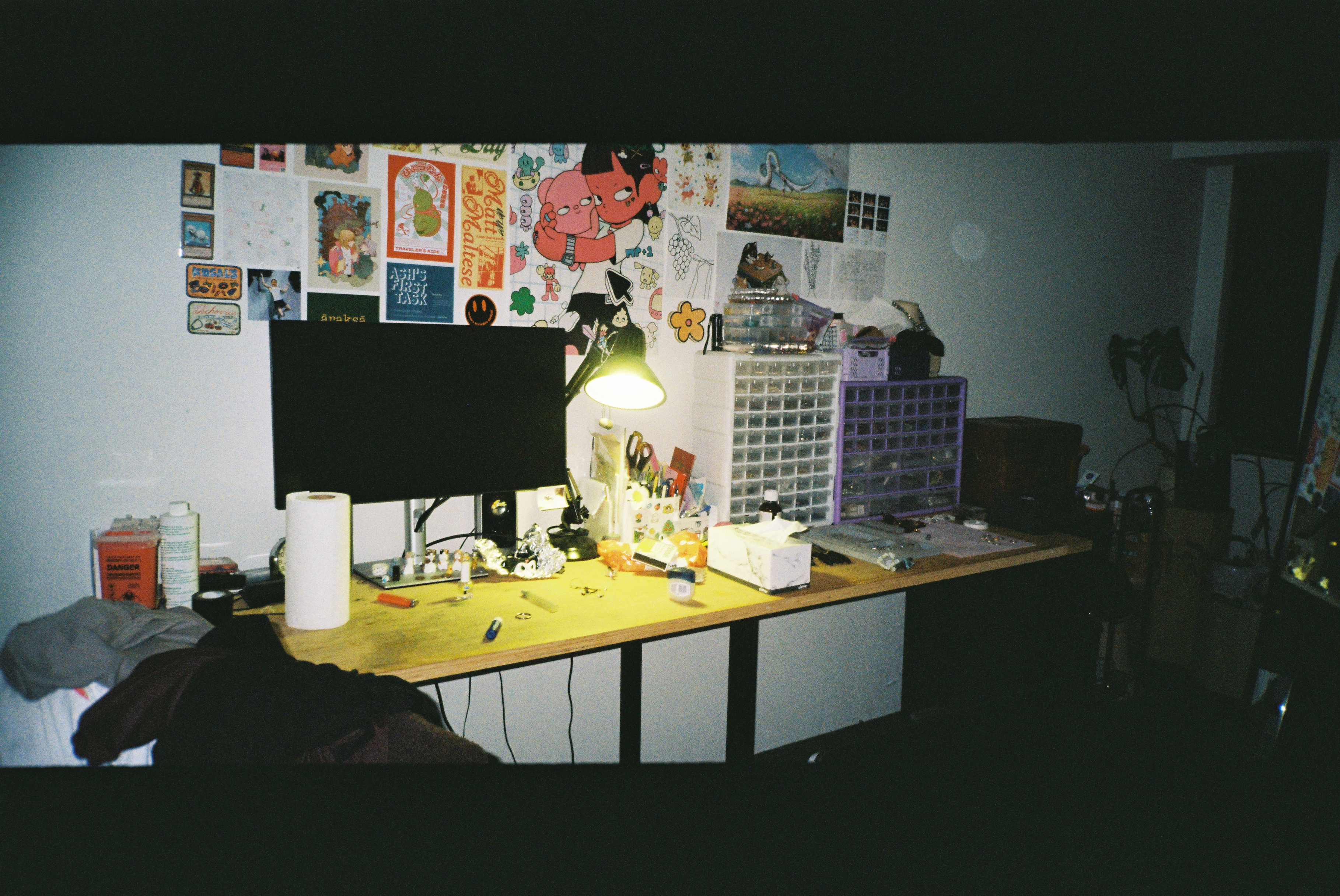 A desk lit by a single lamp, walls covered in art and prints.